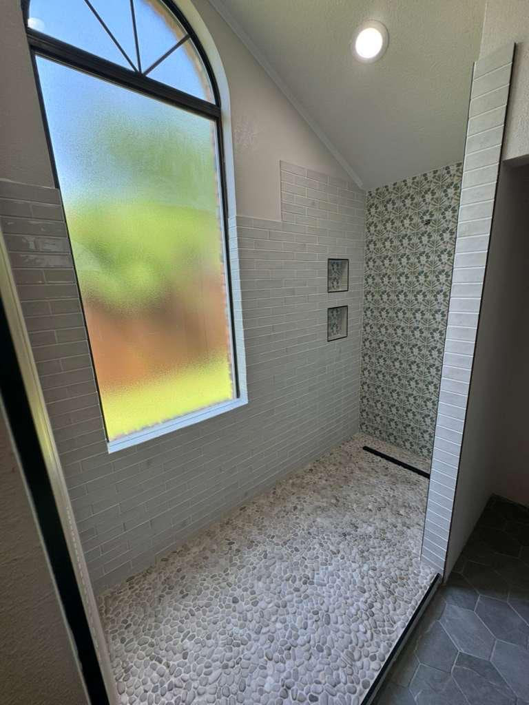 Bright walk-in shower with white subway-style walls, a patterned accent wall, pebble floor and tall frosted window